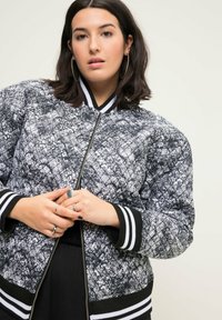 Prošívaná bomber bunda v černé a bílé barvě s texturovaným květinovým vzorem, s předním zipem a proužkovanými žebrovanými manžetami a límcem.