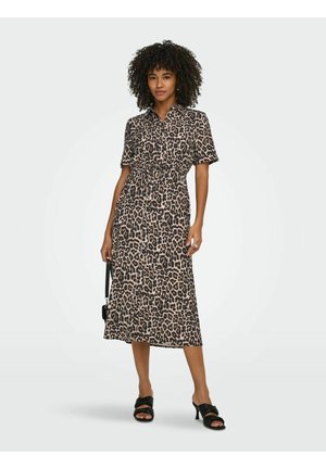 Nainen kiharat hiukset leopardikuvioisessa midimekossa, mustissa korkokengissä, pitelee pientä mustaa käsilaukkua, seisoo yksivärisen taustan edessä.