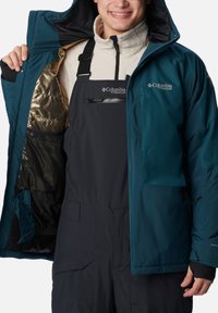 Türkisfarbene isolierte Jacke mit Reißverschluss, Netzfutter und einer großen Tasche. Trägt man über dunklen Latzhosen mit verstellbaren Trägern.