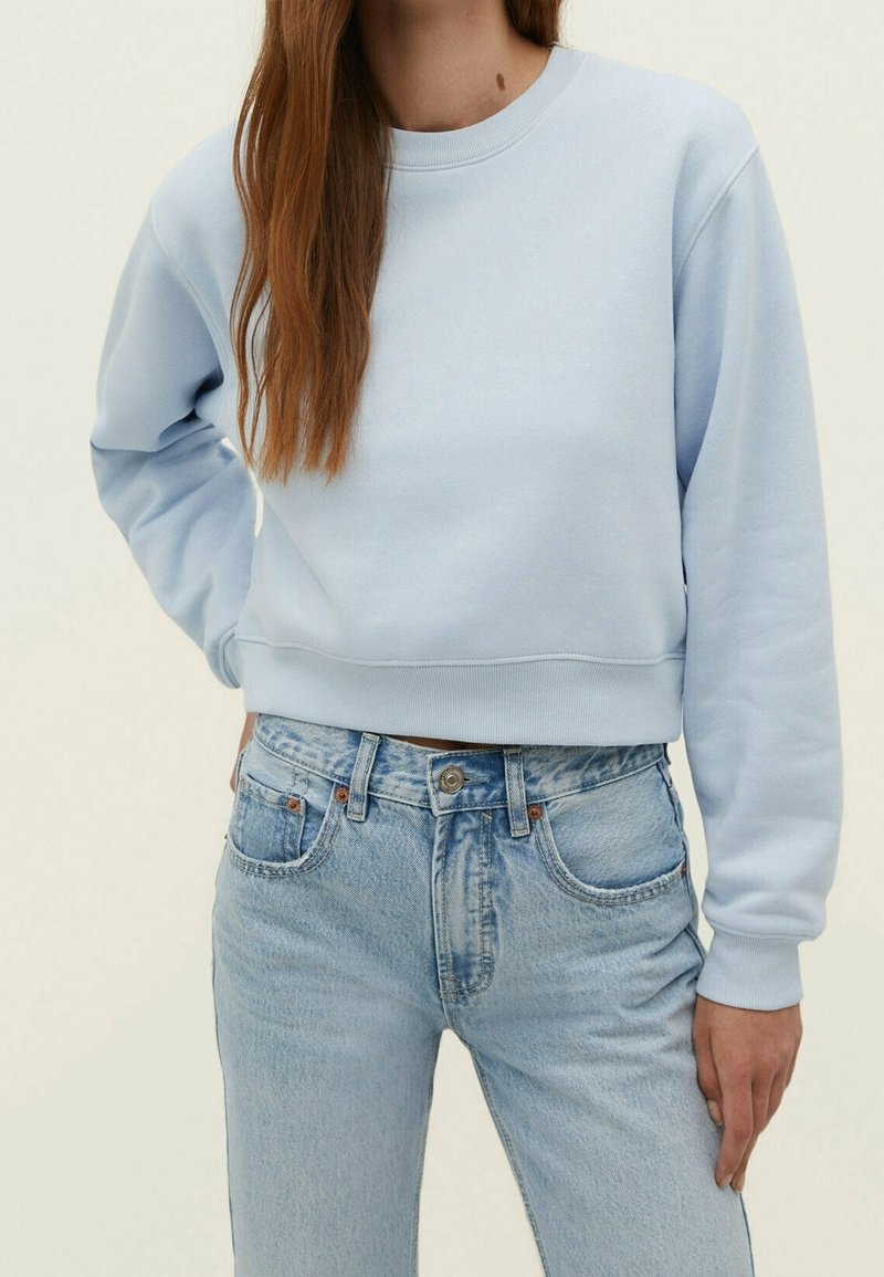 Sweatshirt court bleu clair avec des manches longues, un col rond et des poignets côtelés, associé à un jean taille haute bleu délavé.