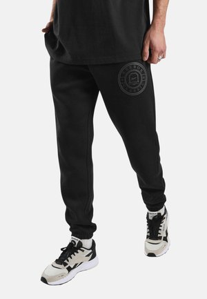 Persoon die zwarte Reebok joggingbroek draagt met een rond logo op de dij en witte en zwarte Reebok sneakers, staand tegen een effen achtergrond.