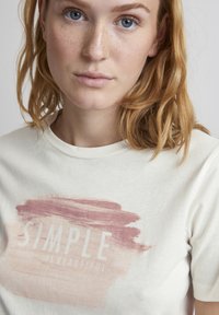 Creme-kleurig katoenen T-shirt met een roze penseelstreek ontwerp en de tekst "SIMPLE IS BEAUTIFUL" in het wit. Korte mouwen, ronde hals.