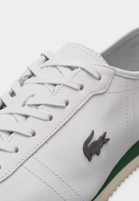 Λευκό δερμάτινο αθλητικό παπούτσι με λευκά κορδόνια, λεπτομερείς ραφές και ένα μικρό σκοτεινό λογότυπο κροκοδείλου στην πλευρά πάνω από μια πράσινη ρίγα.