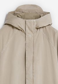 Chaqueta con capucha de un color beige claro, fabricada con un tejido liso y resistente al agua. Presenta un cierre de cremallera en la parte delantera y mangas raglán con detalles de costura sutiles.
