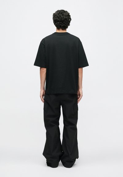 Μαύρο βαμβακερό T-shirt σε oversized γραμμή με κοντά μανίκια και στρογγυλή λαιμόκοψη, σε συνδυασμό με χαλαρά μαύρα cargo παντελόνια με πλαϊνές τσέπες.