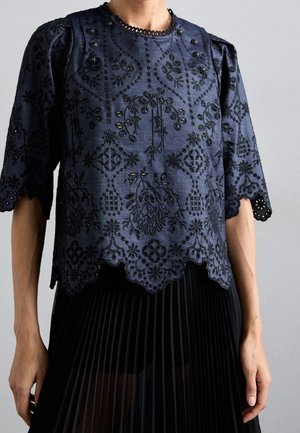 Granatowa bluzka z czarną kwiatową haftem, falistymi krawędziami, bufkami na rękawach i ozdobnymi guzikami, zestawiona z czarną plisowaną spódnicą.