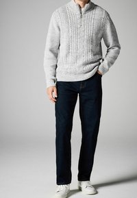 Grauer, mit Kabelmuster gestrickter Pullover mit halbem Reißverschluss, kombiniert mit dunkelblauen Jeans und weißen Turnschuhen. Der Pullover hat strukturierte Muster und gerippte Bündchen.
