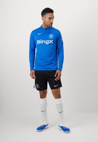 Blå langærmet sportsbluse med høj hals, hvid "Bingx" logo, kombineret med sorte shorts med logo, hvide sokker og blå fodboldstøvler.