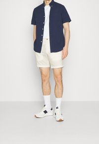 Námořnická košile s krátkým rukávem na knoflíky, bílý tričko s kulatým výstřihem, béžové šortky, bíl é ponožky a bílé tenisky s námořnickými akcenty.