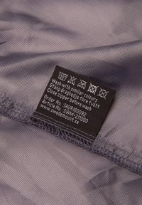 Tessuto grigio con un delicato motivo a griglia, con un'etichetta di cura nera che mostra le istruzioni di lavaggio, il numero d'ordine e il sito web.