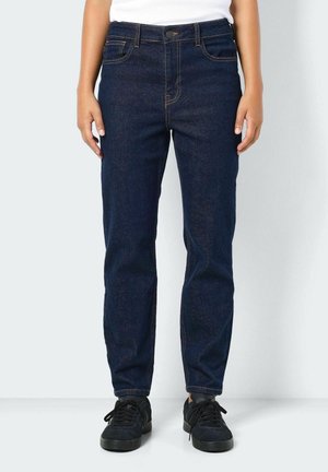 Personne portant un jean droit bleu foncé et des baskets noires, debout les bras détendus le long du corps.
