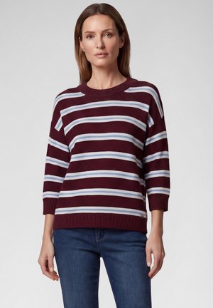 Woman standing wearing a maroon sweater with white and blue horizontal stripes and blue jeans against a plain background.