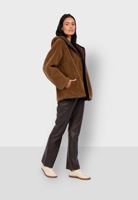 Braune Fleecejacke mit Kapuze, großen Taschen und lockerer Passform, kombiniert mit braunen Lederhosen und hellen Sneakers.