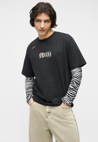 Νεαρός με σκούρα μαλλιά που φοράει μαύρο μπλουζάκι Nirvana πάνω από μακρυμάνικο ζεβροπατινισμένο και μπεζ παντελόνια, με τα χέρια στις τσέπες, ουδέτερο φόντο.