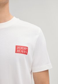 Biała bawełniana koszulka z krótkim rękawem, z czerwonym prostokątnym logo na lewej klatce piersiowej, które głosi "JOOP! JEANS."