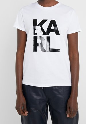 Person trägt ein weißes T-Shirt mit kurzen Ärmeln, auf dem in Schwarz "KARL" steht und ein stilisiertes Profilbild zu sehen ist, kombiniert mit schwarzen Lederhosen.