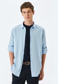 Lyseblå langærmet skjorte i et blødt stof med knaplukning fortil, rullede ærmer og en afslappet pasform over en mørk T-shirt med rund hals.