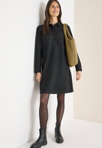 Vestido negro de manga larga con tela texturizada, con cuello de solapa, tapeta de botones y dos bolsillos en el pecho. Combinado con botas negras gruesas.