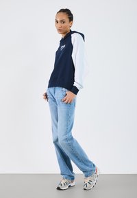Felpa navy con maniche bianche e azzurro chiaro, con logo frontale. Abbinata a jeans azzurro chiaro e sneakers beige con dettagli neri.