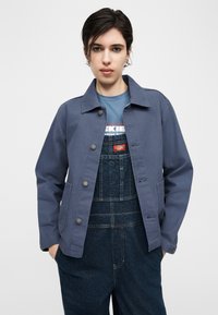 Άτομο με κοντά σκούρα μαλλιά φοράει μπλε μπουφάν πάνω από μπλε μπλούζα Dickies και σκούρα τζιν φορμάκια, τα χέρια στις τσέπες, ουδέτερο φόντο.