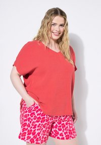 Rød struktureret V-hals top parret med lyserøde shorts med et iøjnefaldende leopardmønster. Modellen står med den ene hånd i lommen og smiler.