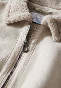 Beige Fleecejacke mit einem weichen, strukturierten Kragen. Mit einem silbernen Reißverschluss und hellen Nähten. Etikett im Kragen sichtbar.