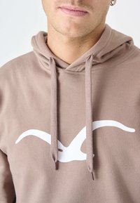 Felpa marrone realizzata in un tessuto morbido con un logo bianco sul davanti che presenta un design simile a un uccello. Ha un cappuccio con cordoncino e dettagli a coste.