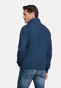 Marineblauwe zip-up jas met een gestructureerd oppervlak, hoge kraag en elastische manchetten. G gedragen met blauwe jeans, wat een eigentijdse uitstraling toont.