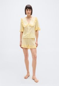 Femme debout pieds nus portant un ensemble de pyjama en coton jaune pâle avec des bordures en dentelle œillet sur le col, les manches et le short, sur un fond blanc.