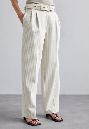 Pantalon à rayures blanches avec taille haute, devant plissé et ceinture assortie. Le matériau a une texture lisse. Associé à des talons noirs à lanières.