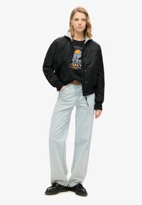 Veste bomber noire avec une capuche grise, portée sur un sweat à graphic, associée à un jean large bleu clair et des chaussures noires.