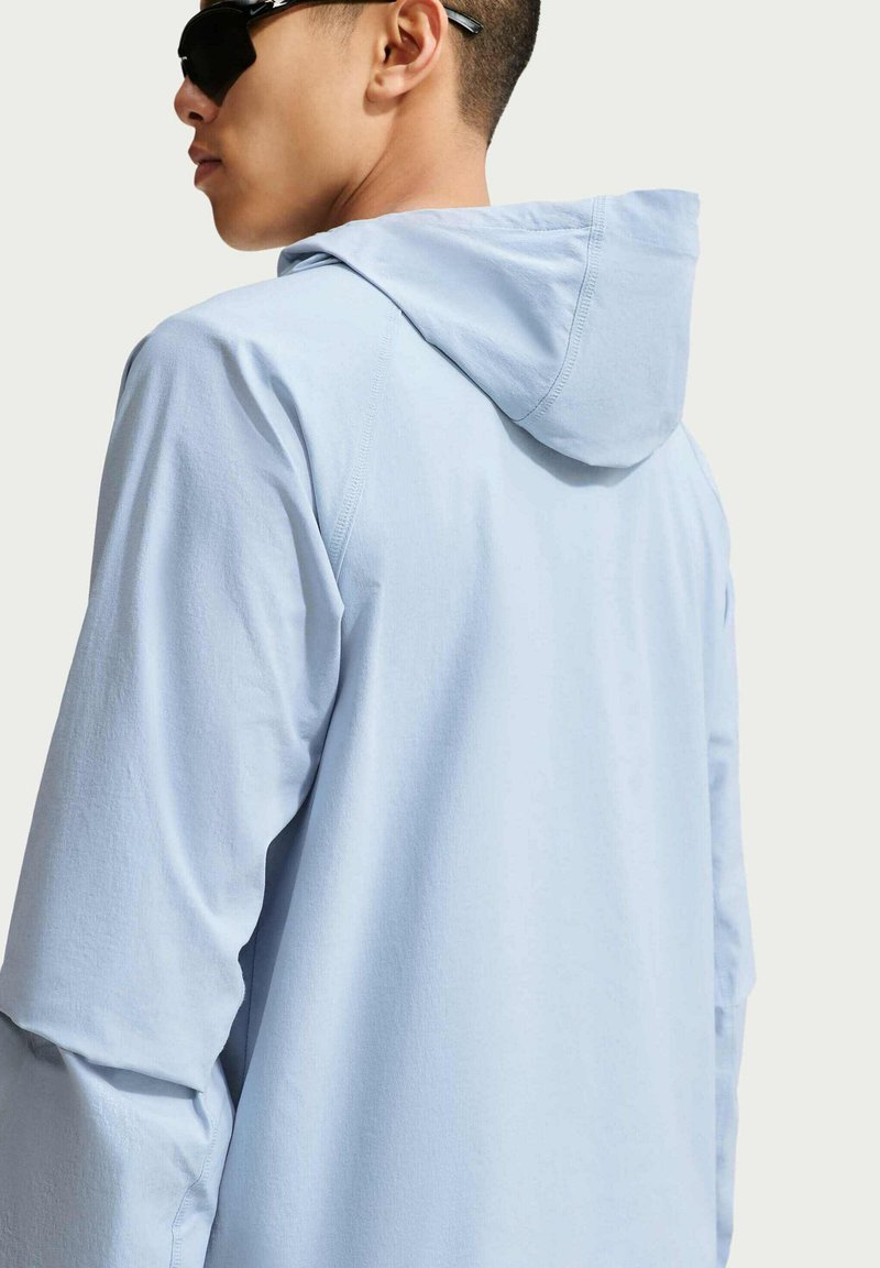 Junger Mann mit schwarzen Sonnenbrillen und lockerem hellblauen Hoodie, der mit dem Rücken zu einem schlichten hellen Hintergrund steht.