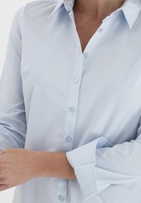 Kobieta poprawiająca mankiety jasnoniebieskiej koszuli z kołnierzykiem, widoczna od szyi do talii na białym tle.