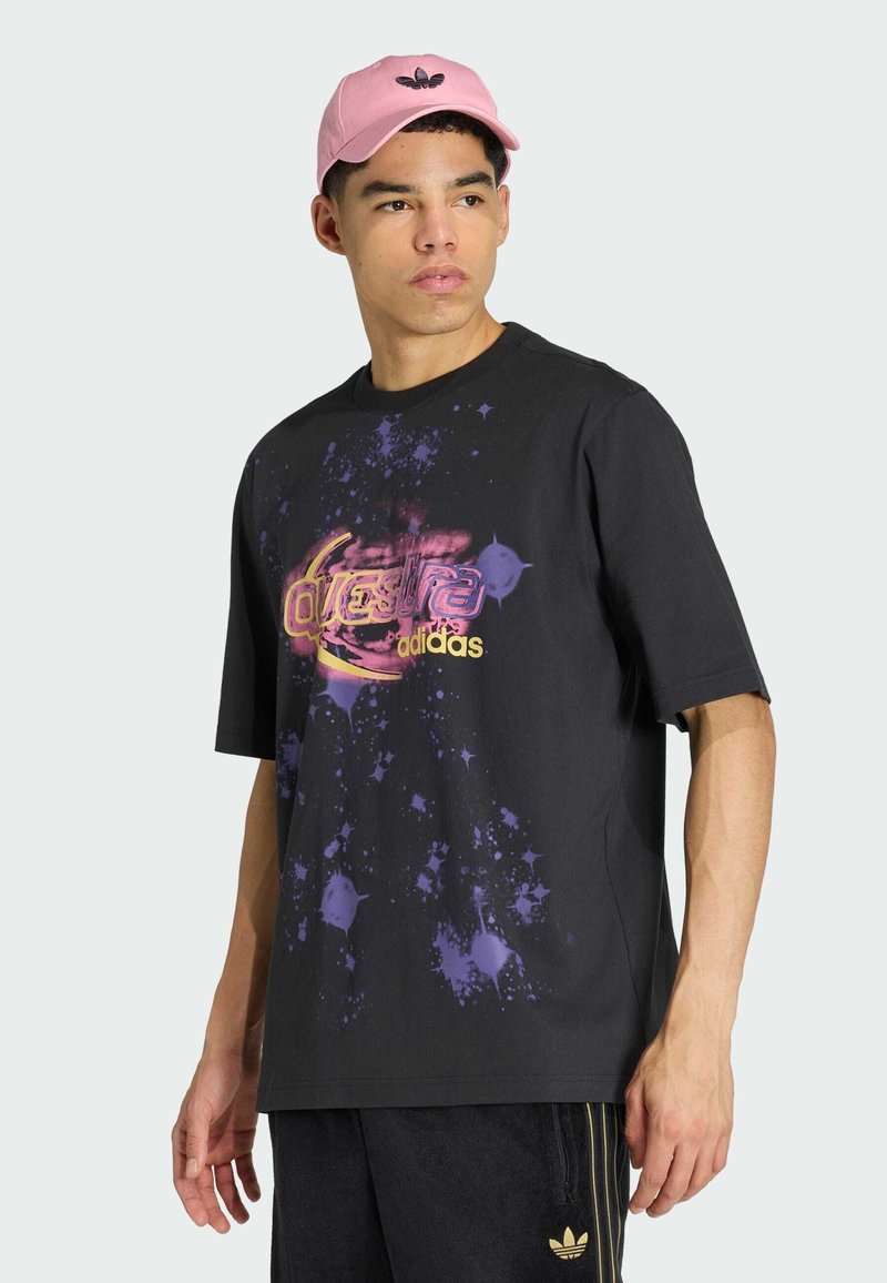 Junger Mann trägt ein schwarzes Adidas "Questra" Grafik-T-Shirt, schwarze Adidas-Hose mit goldenen Streifen und eine pinke Adidas-Kappe, schaut zur Seite.