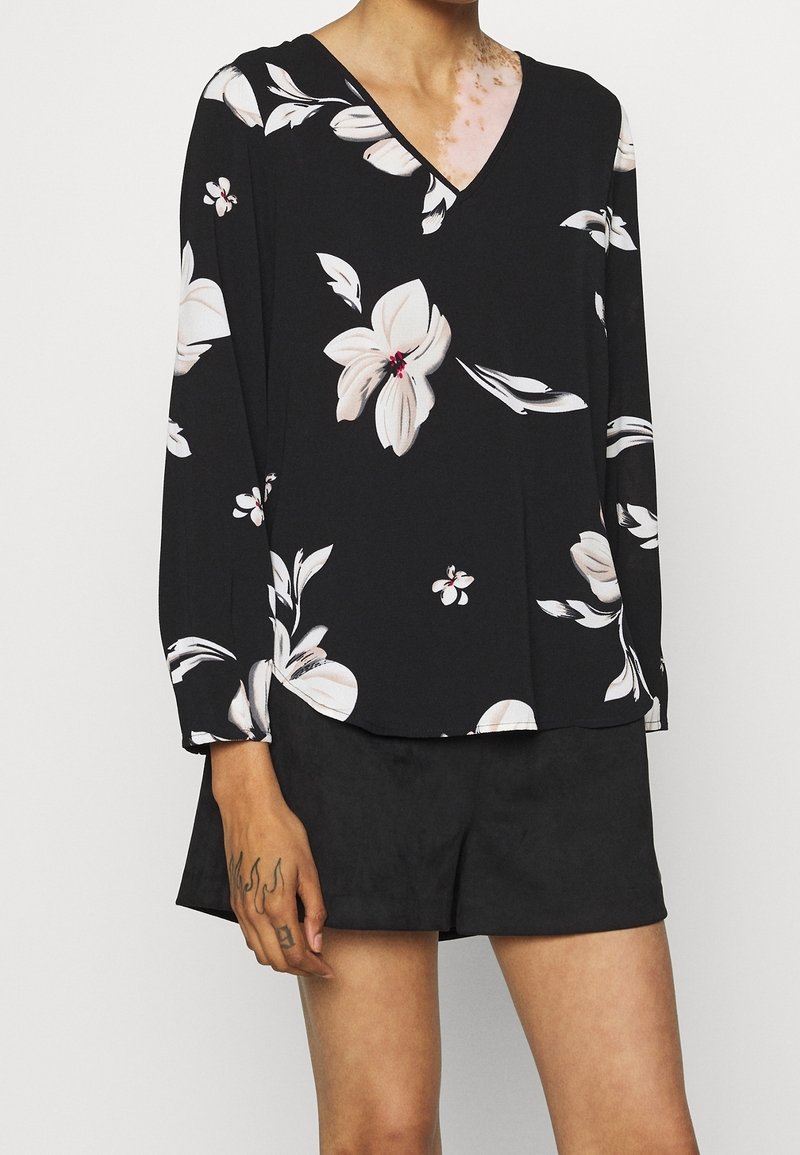 Femme portant un chemisier noir à manches longues avec un grand imprimé floral blanc et un short noir, montrant une main tatouée et une partie du cou.