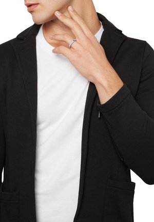 Cárdigan negro de corte ajustado y solapas de pico, superpuesto a una camiseta blanca. Se puede ver un anillo plateado en la mano del modelo.