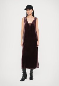 Un vestido de terciopelo burdeos oscuro con un escote en V pronunciado, aberturas laterales y un forro transparente. Combinado con botas negras hasta la rodilla y una gorra.