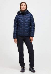 Marineblauwe gewatteerde jas met een golvend doorgestikt ontwerp, ritssluiting aan de voorkant en capuchon. Draagt zwart broek en wandelschoenen.