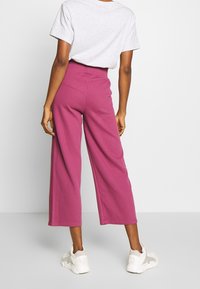 Wide-leg, ribbed trousers in magenta, made from soft fabric. They feature a high waist and are paired with white sneakers.