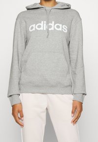 Felpa grigia realizzata in tessuto morbido, con tasca frontale e logo "adidas" bianco. Il design include un cappuccio regolabile con cordino e polsini a coste.