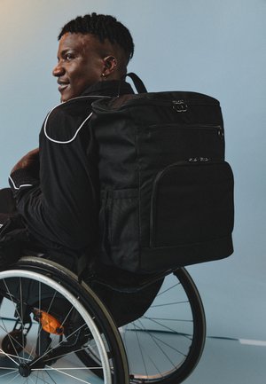 Melnā mugursoma ar vairākiem ar rāvējslēdzējiem aizveramajiem kabatām, izgatavota no izturīga materiāla, ar strukturētu dizainu un gludu tekstūru, valkāta uz ratiņkrēsla.