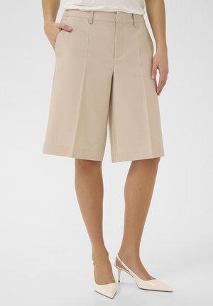 Pantaloni corti beige su misura con taglio dritto, dotati di tasche laterali, passanti per cintura e una texture liscia, abbinati a décolleté a punta.