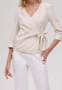 Béžovo-krémový pruhovaný top s obvazem a tříčtvrtečními rukávy, se zavazováním v pase, spárovaný s bílými kalhotami s bočními knoflíky.