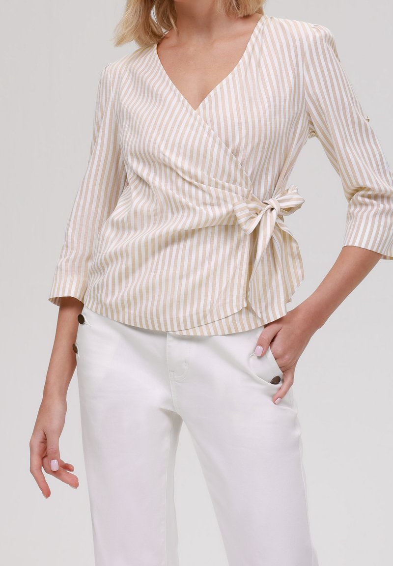 Béžovo-krémový pruhovaný top s obvazem a tříčtvrtečními rukávy, se zavazováním v pase, spárovaný s bílými kalhotami s bočními knoflíky.