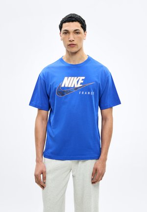 Giovane uomo con capelli corti e ricci che indossa una maglietta blu Nike Francia e pantaloni chiari su uno sfondo bianco uniforme.