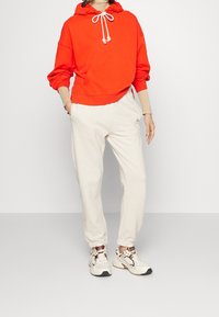 Felpa rossa con cappuccio e cordini, tasca a canguro, pantaloni della tuta beige con polsini elastici e sneakers bianche con dettagli marroni.