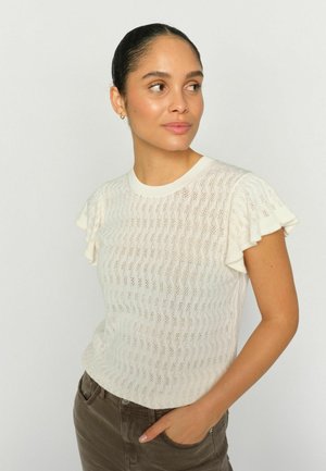 Femeie cu părul prins în coc, purtând un top alb texturat cu mâneci scurte și pantaloni maro, pozând cu o mână în buzunar și privind spre lateral.