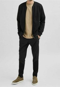 Černá prošívaná bomber bunda přes béžové tričko, doplněná černými zúženými kalhotami a hnědými teniskami. Jednoduchý design a tlumené barvy.