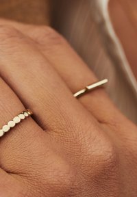 Zwei goldene Ringe an einer Hand: Einer zeigt eine Reihe kleiner, polierter Scheiben; der andere hat ein schlankes, stabförmiges Design. Glatte Oberfläche.