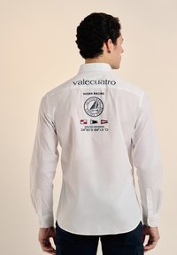 Camisa blanca de manga larga con cuello, logotipo "valecuatro" y gráficos de temática náutica en la parte posterior. Textura suave con puños abotonados.